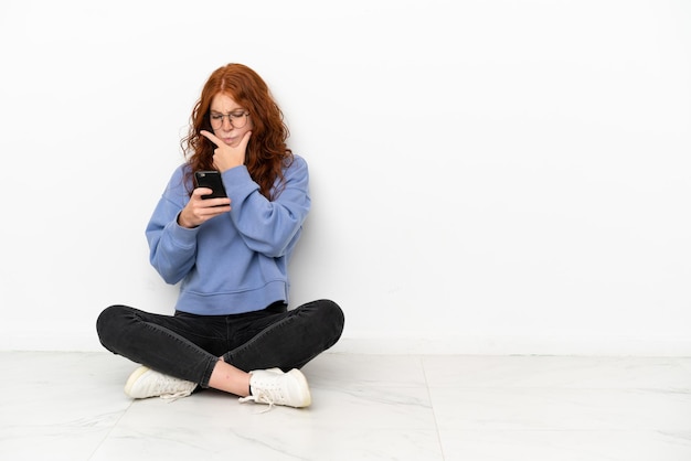 Ragazza dai capelli rossi adolescente seduta sul pavimento isolata su sfondo bianco pensando e inviando un messaggio