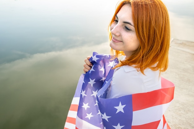 Ragazza dai capelli rossi sorridente felice con la bandiera nazionale degli Stati Uniti sulle sue spalle. Giovane donna positiva che celebra il giorno dell'indipendenza degli Stati Uniti.