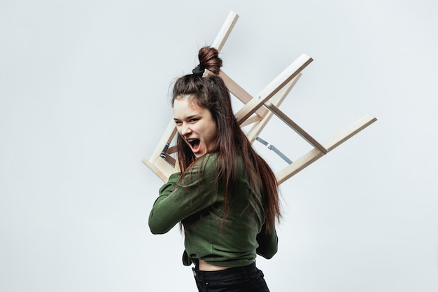 Ragazza dai capelli scuri alla moda aggressiva con un'acconciatura vestita in camicia verde, pantaloncini neri, collant e scarpe è in posa con lo sgabello di legno sullo sfondo bianco in studio.