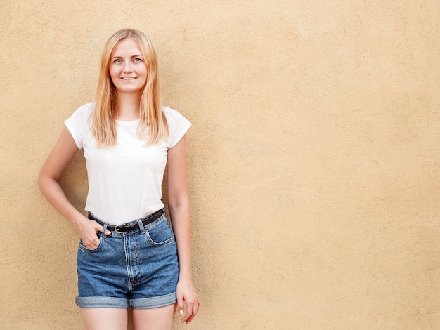 Ragazza dei pantaloni a vita bassa che indossa maglietta bianca in bianco e jeans che posano contro il muro di strada ruvido, stile minimalista dell'abbigliamento urbano