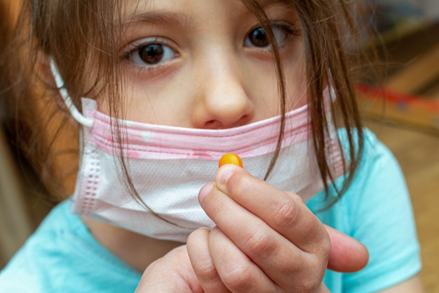 Ragazza del bambino che indossa maschera protettiva medica con una pillola in una mano a casa durante l'isolamento di quarantena