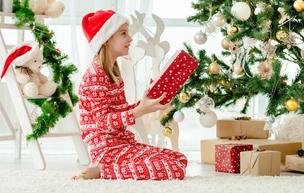 Ragazza del bambino che tiene nel contenitore di regalo rosso di tempo di Natale a casa. Bambino che festeggia il nuovo anno con regali e albero di Natale