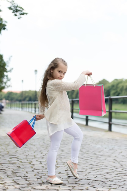 Ragazza del bambino con i capelli lunghi che ama lo shopping Ragazza fashionista che fa shopping con borse rosa Concetto di shopping Alla ragazza piace comprare vestiti Ragazza che fa shopping sul viso calmo porta borse della spesa sullo sfondo urbano