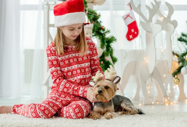 Ragazza del bambino con il cane nel periodo natalizio a casa con decorazioni natalizie. Bambino che indossa un costume rosso e un cagnolino in camera con decorazioni di Capodanno
