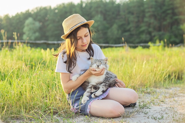 Ragazza del bambino in cappello con gatto lanuginoso grigio tra le braccia. Bellissimo sfondo del paesaggio di campagna al tramonto.