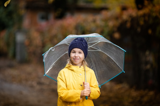 Ragazza del bambino in giacca gialla e cappello blu scuro con l'ombrello trasparente nel parco di autunno