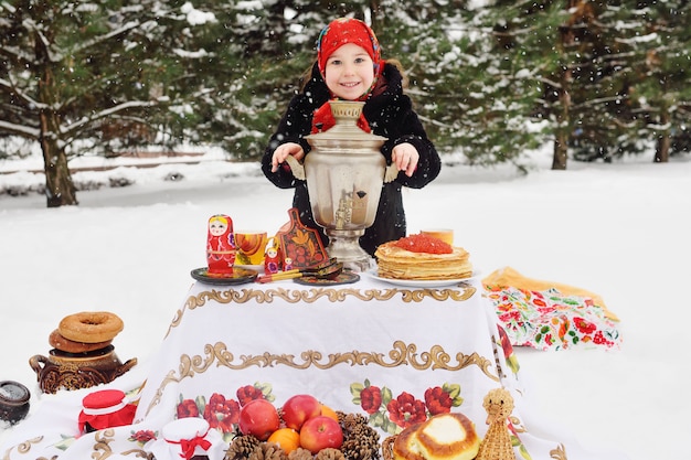 Ragazza del bambino in una pelliccia e in una sciarpa in stile russo con in mano una grande samovar