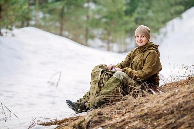 Ragazza del cacciatore che si siede con lo zaino e la foresta di neve all'aperto di acqua potabile sullo sfondo