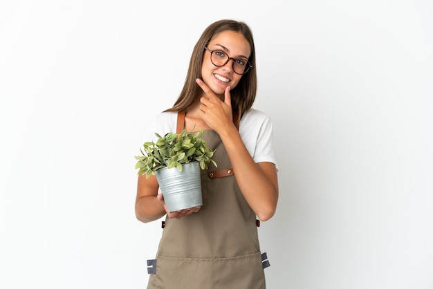 Ragazza del giardiniere che tiene una pianta sopra la parete bianca isolata felice e sorridente