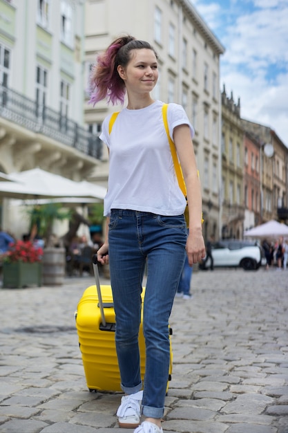 Ragazza del viaggiatore che cammina con lo zaino e la valigia gialla lungo la strada della città turistica