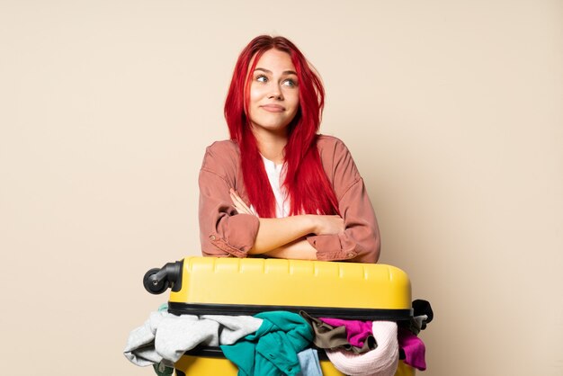 Ragazza del viaggiatore con una valigia piena di vestiti isolati su fondo beige che fa gesto di dubbi mentre solleva le spalle