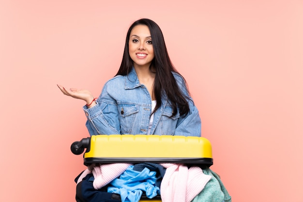 Ragazza del viaggiatore con una valigia piena di vestiti sopra la parete rosa che tiene immaginario spazio vuoto sul palmo