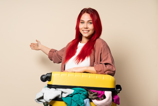 Ragazza del viaggiatore con una valigia piena di vestiti sulla parete beige che estende le mani al lato per invitare a venire