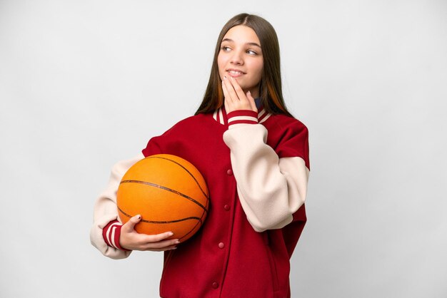 Ragazza dell'adolescente che gioca a basket su sfondo bianco isolato alzando lo sguardo mentre sorride