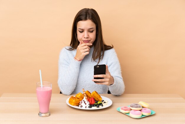 Ragazza dell'adolescente che mangia le cialde isolate sulla parete beige che pensa e che invia un messaggio