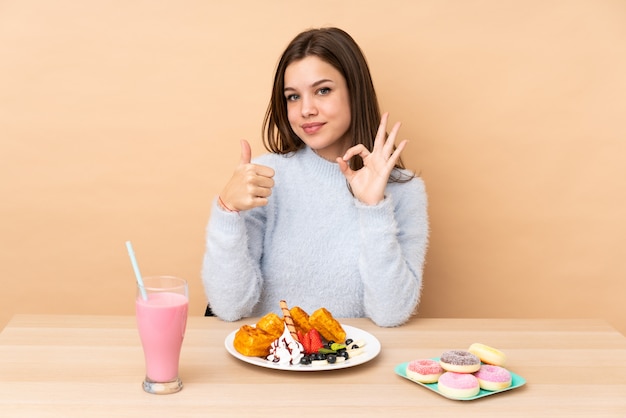 Ragazza dell'adolescente che mangia le cialde sulla parete beige che mostra segno e pollice giusti sul gesto