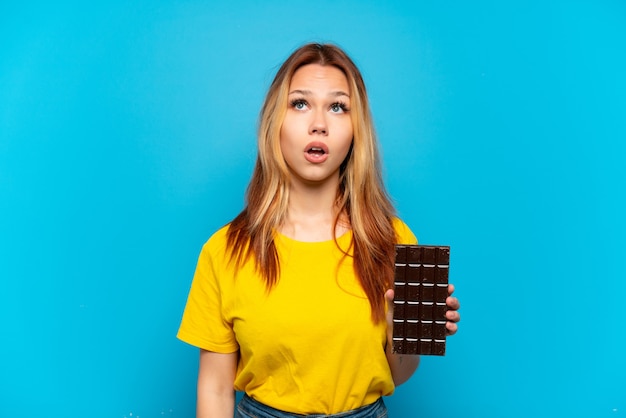 Ragazza dell'adolescente che tiene chocolat sopra fondo blu isolato che osserva in su e con l'espressione sorpresa