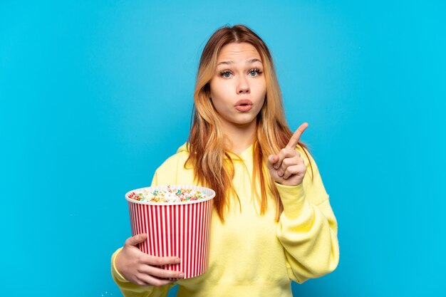Ragazza dell'adolescente che tiene i popcorn sopra fondo blu isolato che intende realizzare la soluzione mentre solleva un dito