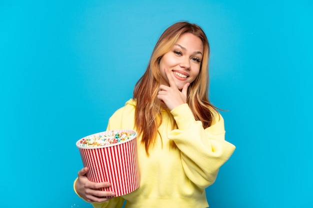 Ragazza dell'adolescente che tiene i popcorn sopra fondo blu isolato felice e sorridente