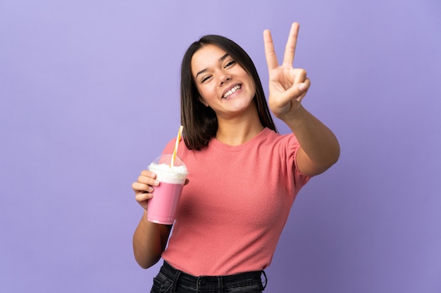 Ragazza dell'adolescente che tiene un milkshake della fragola che sorride e che mostra il segno di vittoria