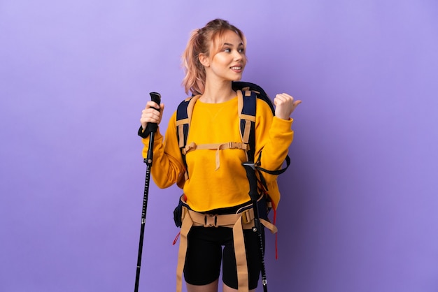 Ragazza dell'adolescente con lo zaino e bastoncini da trekking sopra la parete viola isolata che indica al lato per presentare un prodotto