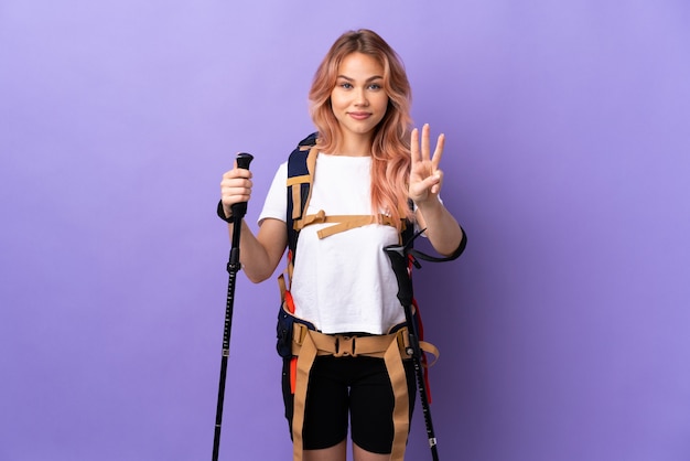 Ragazza dell'adolescente con lo zaino e bastoncini da trekking sopra la porpora isolata felice e che conta tre con le dita