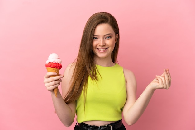 Ragazza dell'adolescente con un gelato della cornetta sopra fondo rosa isolato che fa il gesto dei soldi