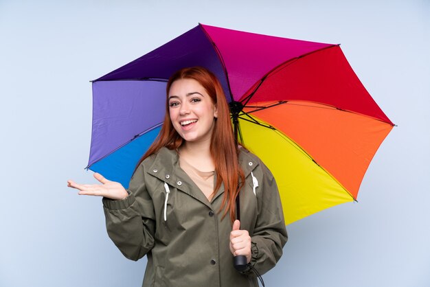 Ragazza dell'adolescente della testarossa che tiene un ombrello sopra il copyspace blu isolato della tenuta della parete immaginario sulla palma