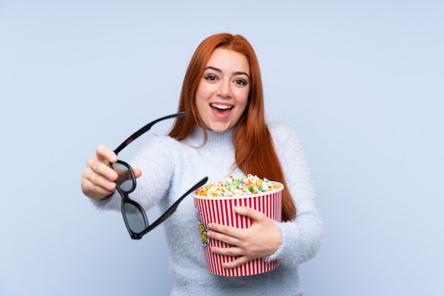 Ragazza dell'adolescente della testarossa sopra l'azzurro con gli occhiali 3d e tenere un grande secchio di popcorn