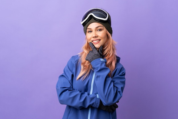 Ragazza dell'adolescente dello sciatore con gli occhiali dello snowboard sopra la parete viola isolata felice e sorridente
