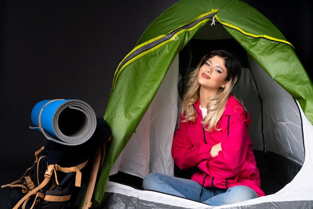 Ragazza dell'adolescente dentro una tenda verde di campeggio sulla parete nera con le armi attraversate e felici