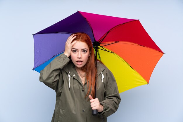 Ragazza dell'adolescente di Redhead che tiene un ombrello sopra il blu con espressione facciale di sorpresa