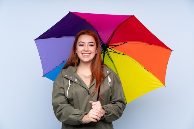 Ragazza dell'adolescente di Redhead che tiene un ombrello sopra il blu con espressione facciale di sorpresa
