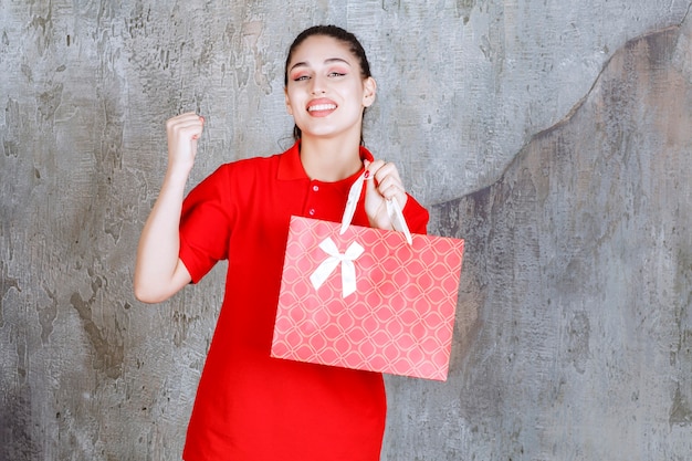 Ragazza dell'adolescente in camicia rossa che tiene una borsa della spesa rossa e che mostra il segno positivo della mano.