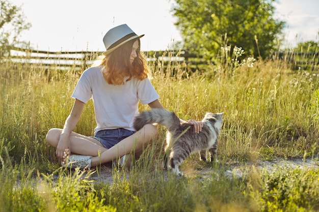 Ragazza dell'adolescente in cappello sulla natura che gioca con il gatto lanuginoso grigio, campagna