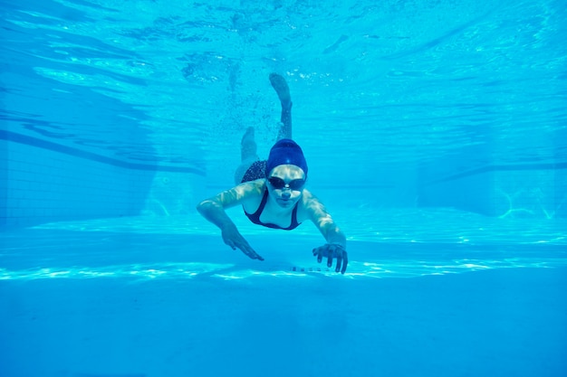 Ragazza dell'adolescente in costume da bagno con gli occhiali di protezione e la cuffia da nuoto che nuota underwater nello stagno