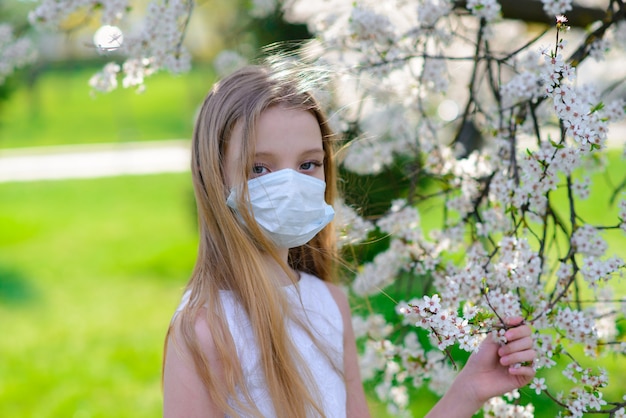 Ragazza dell'adolescente nella mascherina medica in giardino a fioritura primaverile
