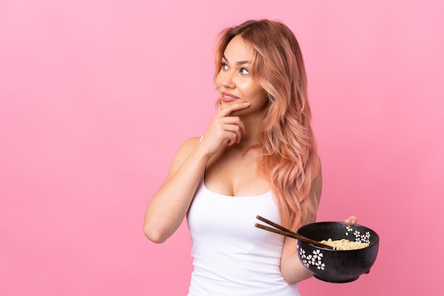 Ragazza dell'adolescente sopra il rosa isolato pensando un'idea e guardando il lato mentre si tiene una ciotola di spaghetti con le bacchette