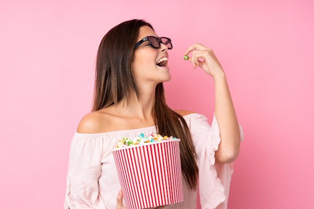 Ragazza dell'adolescente sopra la parete rosa isolata con i vetri 3d e tenere un grande secchio di popcorn