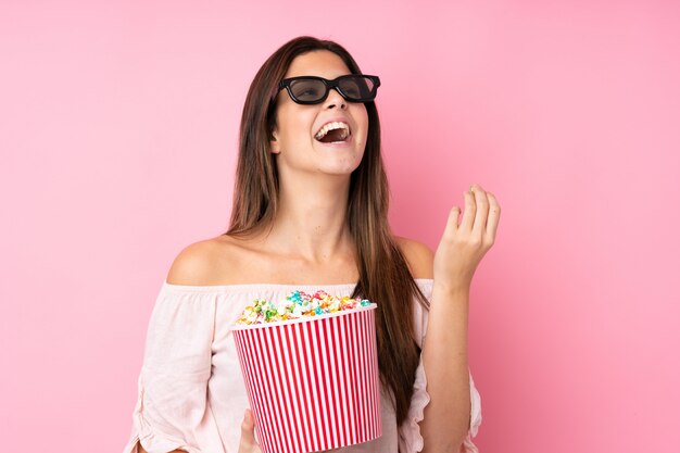 Ragazza dell'adolescente sopra la parete rosa isolata con i vetri 3d e tenere un grande secchio di popcorn