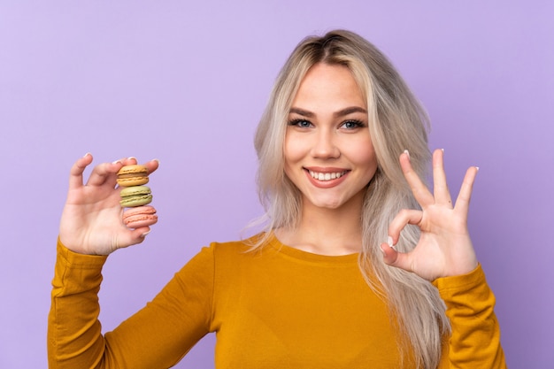 Ragazza dell'adolescente sopra la parete viola che tiene i macarons francesi variopinti e che mostra segno giusto con le dita