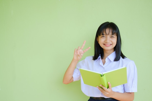 Ragazza dell'allievo del ritratto nella scuola dell'uniforme con il libro verde sulla parete verde. Ragazza asiatica adolescente