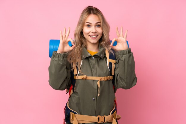 Ragazza dell'alpinista dell'adolescente con un grande zaino isolato sulla parete rosa che mostra un segno giusto con le dita