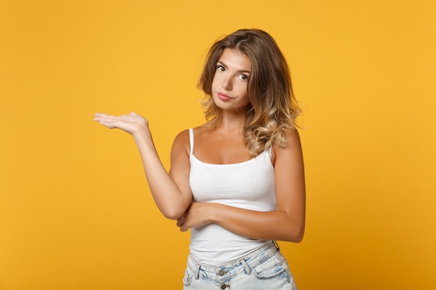 Ragazza della giovane donna in vestiti casuali leggeri che posano isolato sul ritratto dello studio del fondo della parete giallo arancio. Persone sincere emozioni concetto di stile di vita. Mock up spazio di copia. Indicando la mano da parte.