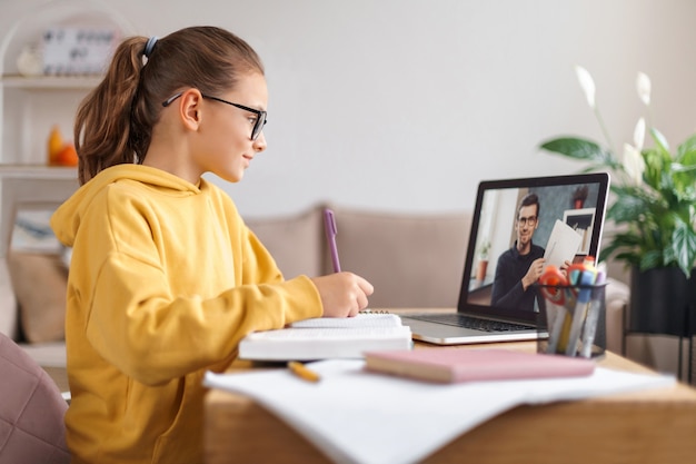 Ragazza della scuola che ha una videoconferenza con l'insegnante online sul laptop nel soggiorno di casa