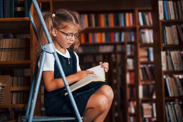 Ragazza della scuola sulla scala in biblioteca piena di libri