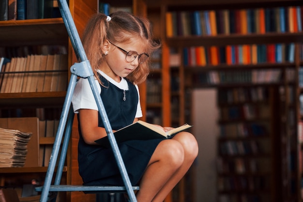 Ragazza della scuola sulla scala in biblioteca piena di libri