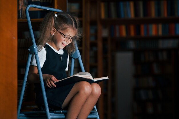 Ragazza della scuola sulla scala in biblioteca piena di libri