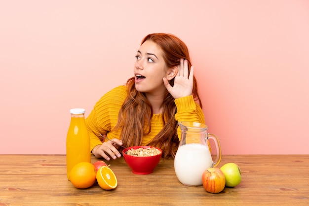 Ragazza della testarossa dell'adolescente che mangia prima colazione in una tabella che ascolta qualcosa