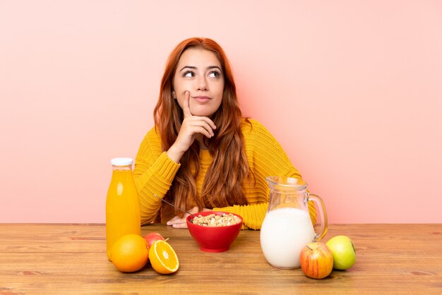 Ragazza della testarossa dell'adolescente che mangia prima colazione in una tavola che pensa un'idea